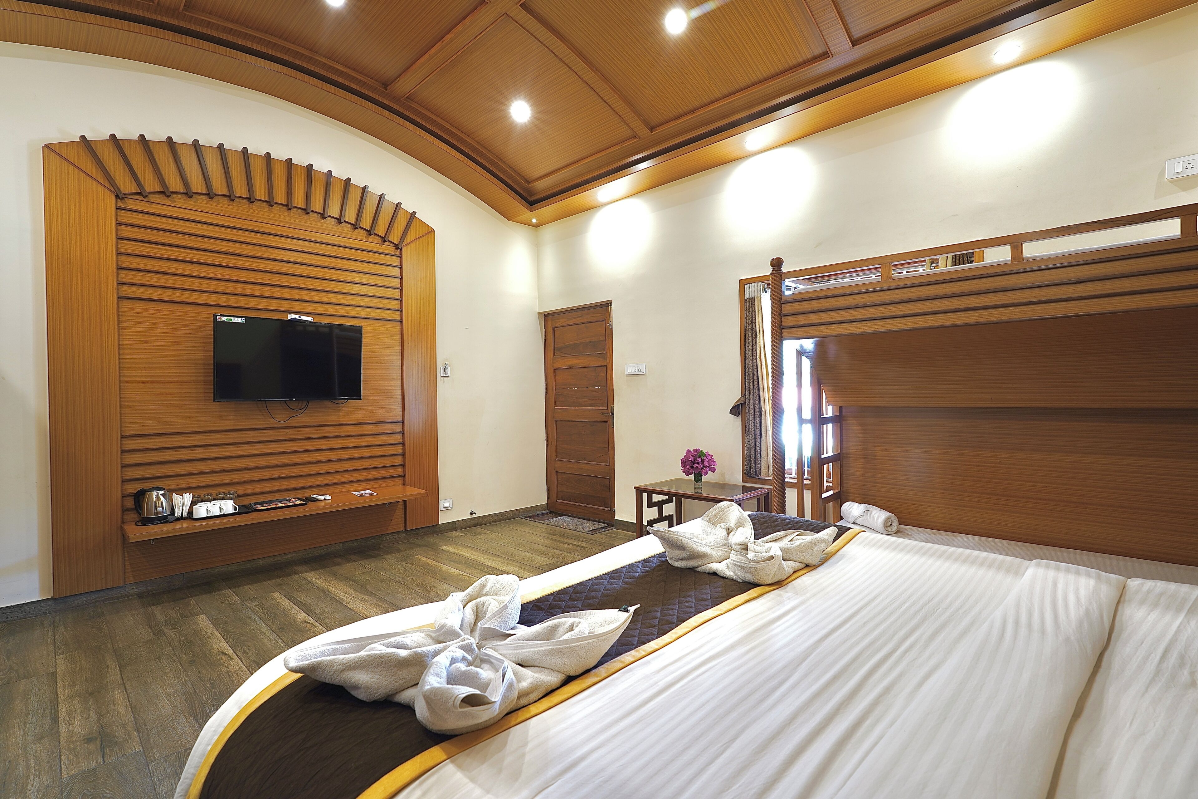 Habitación de hotel con una cama grande, cabecera de madera, un televisor de pantalla plana montado en la pared y una mesita con un jarrón de flores.