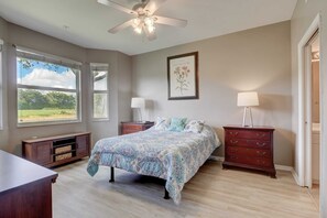 Une chambre à coucher comprenant un lit, deux tables de chevet, une commode, un ventilateur de plafond et une fenêtre donnant sur un paysage verdoyant.