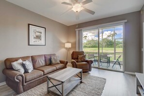 A living room with a brown leather sofa, a wooden coffee table, a floor lamp, and a large sliding glass door leading to an outdoor patio with a table and chairs. - Cypress Woods for Seasonal or Summer Stays (Naples)