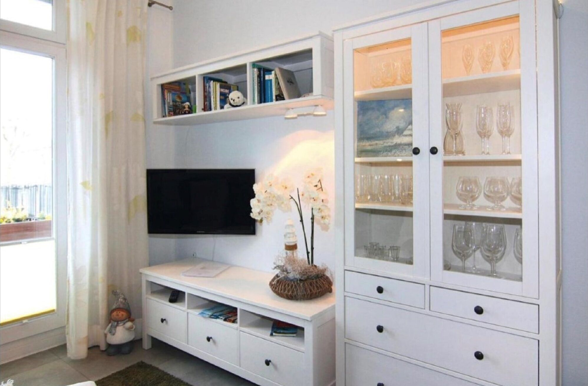 A living room with a flat-screen TV, a white entertainment unit with books and decorative items, a glass cabinet with wine glasses, and a small statue on the floor.