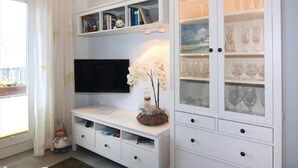 Een woonkamer met een flatscreen-tv, een witte dressoir met boeken en decoratieve objecten, een glazen kast met wijnglazen en een klein beeldje op de vloer.