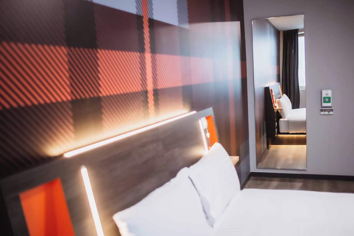 Een hotelkamer met een bed, een nachtkastje, een spiegel en een deur die naar een andere kamer leidt.