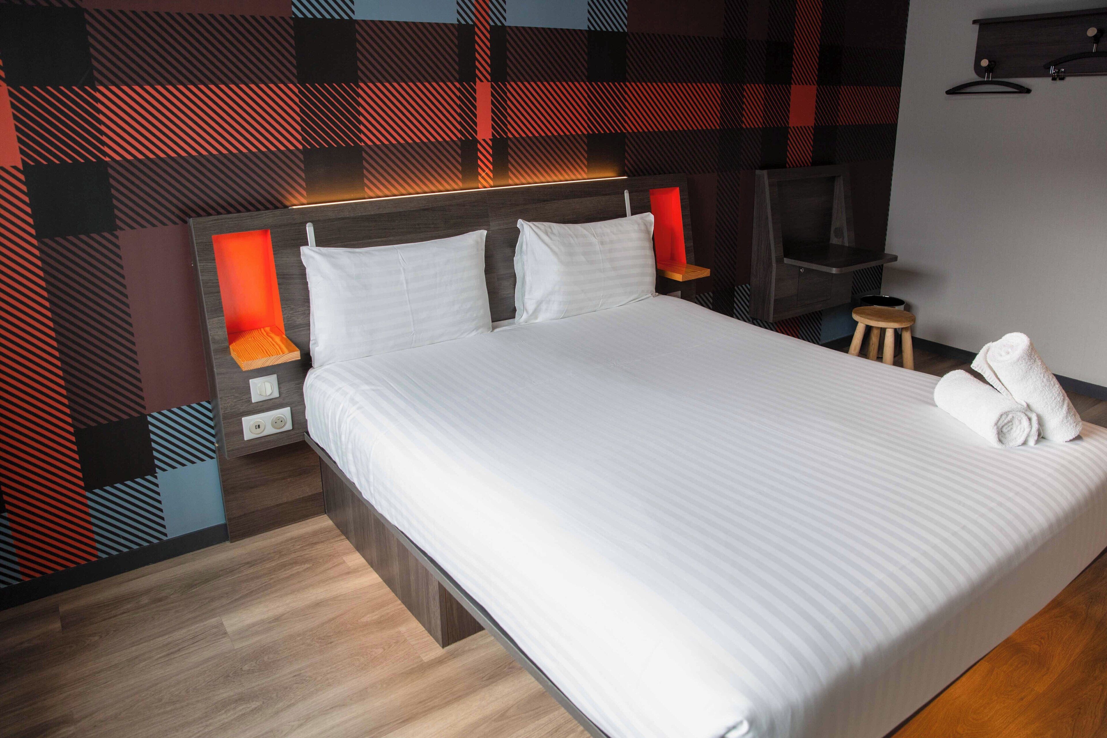 Habitación de hotel moderna con una cama grande, lámparas de noche, un televisor y un taburete pequeño.