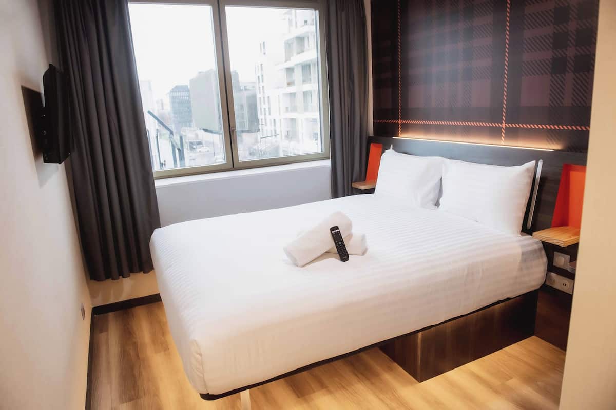 Een hotelkamer met een bed, een aan de muur bevestigde televisie, een raam met gordijnen en uitzicht op de stad.