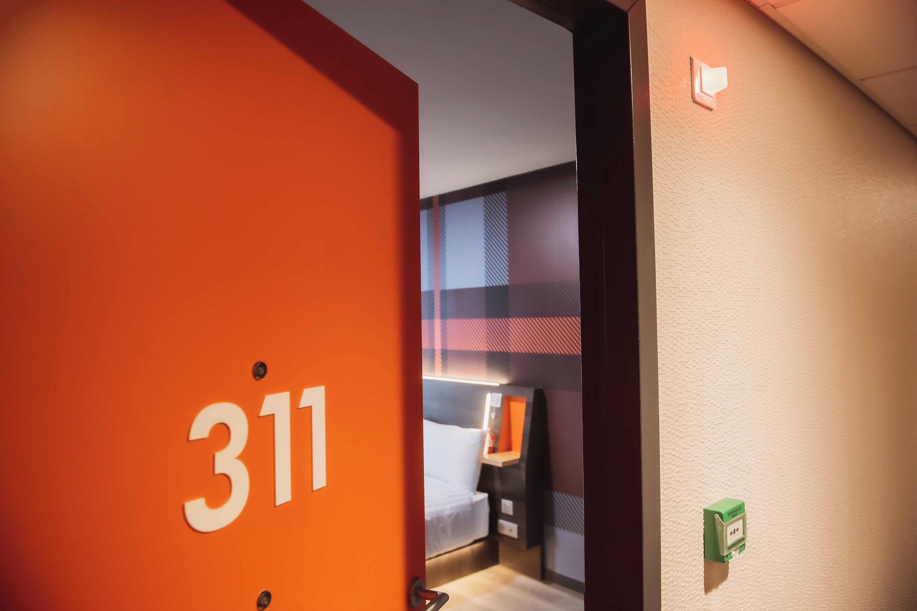 Een hotelkamer met een deur waarop '311' staat, een bed, een nachtkastje en een inbouwschakelaar.
