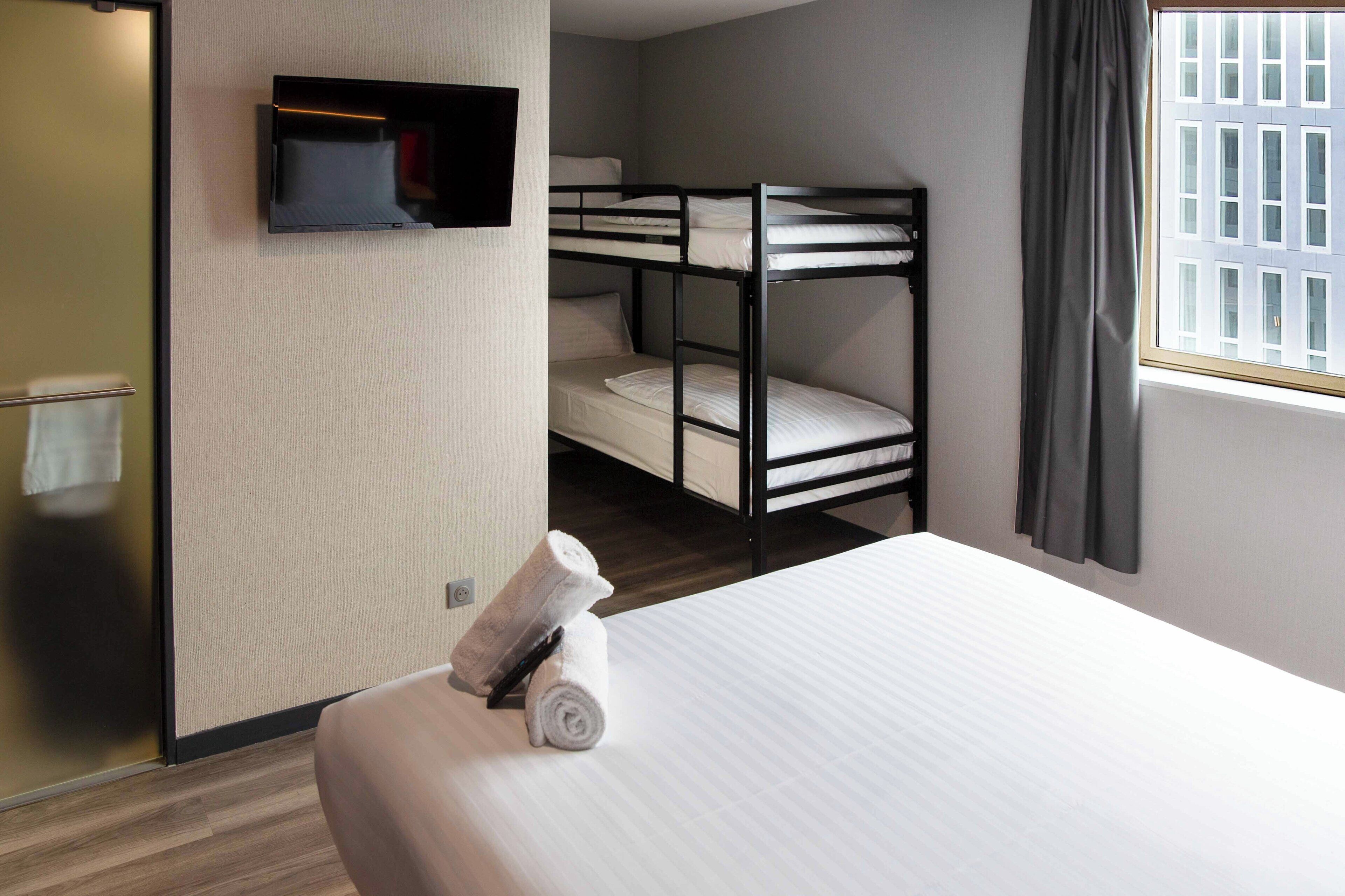 Habitación de hotel con literas, televisor montado en la pared, ventana con cortinas y una cama bien hecha.