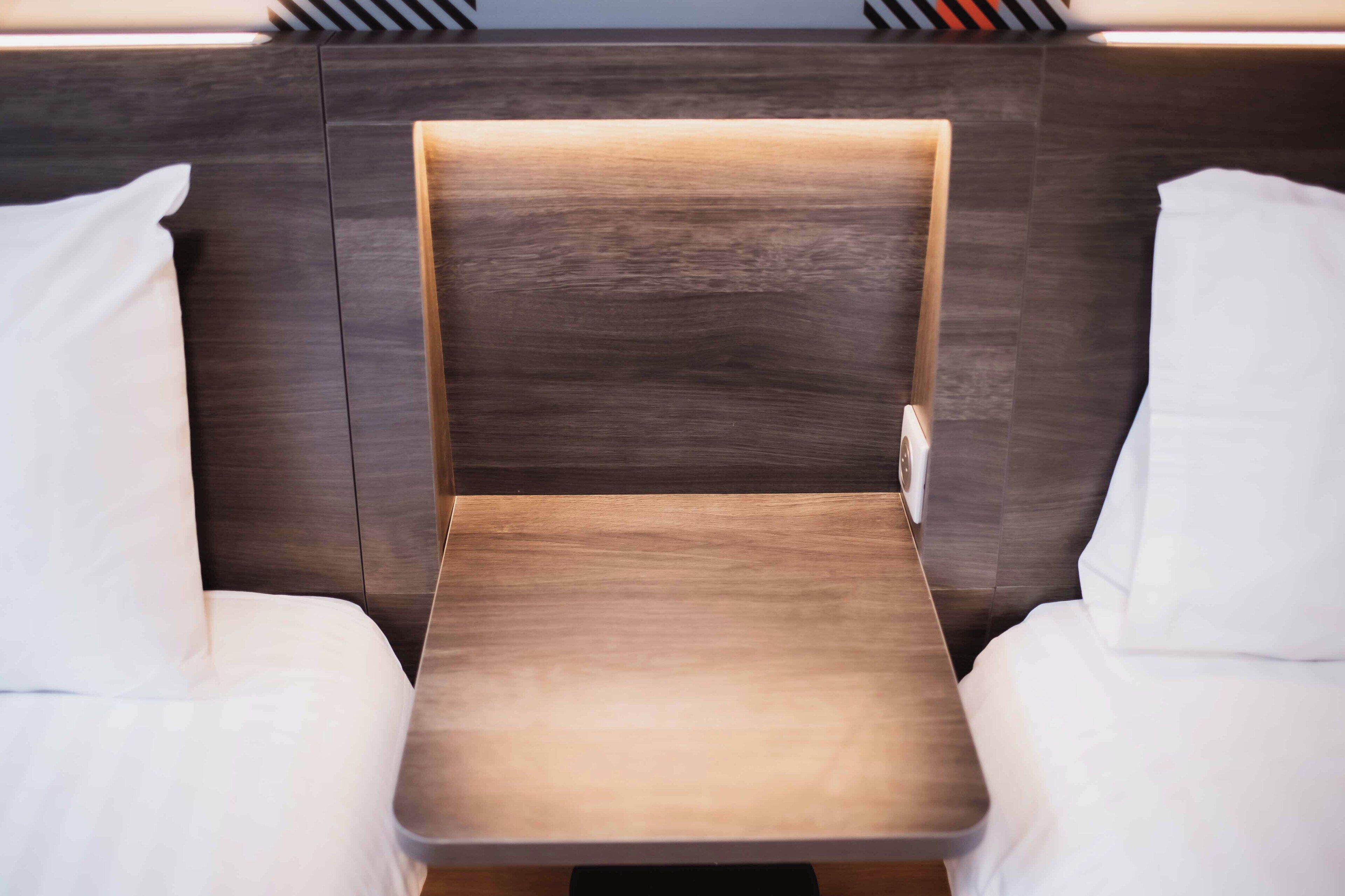 A modern hotel room with a central bed, a small table, and a headboard with built-in lighting.