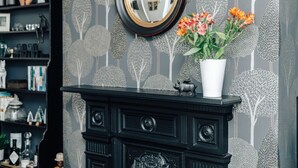 Eine dunkle, hölzerne Kommode mit einem Spiegel darüber, eine weiße Vase mit Blumen und ein Regal mit verschiedenen Gegenständen.