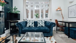 Ein modernes Wohnzimmer mit einem dunkelblauen Sofa, einem Glastisch und einem großen Fenster mit Blick auf Backsteingebäude.