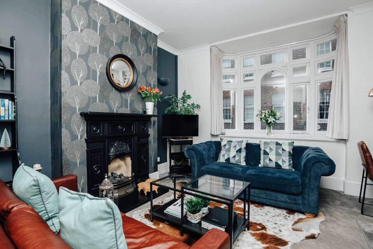 Uma sala de estar com lareira, um sofá azul, uma mesa de centro e uma janela grande com cortinas.