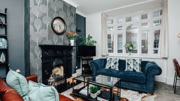 Uma sala de estar com lareira, um sofá azul, uma mesa de centro e uma janela grande com cortinas.