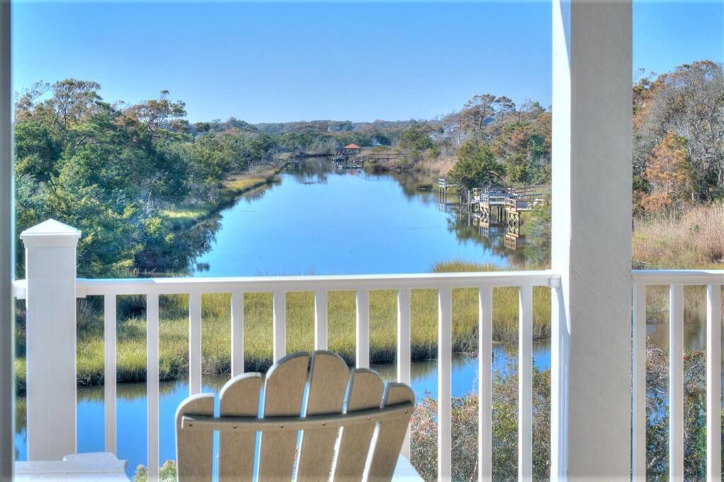 A view from a porch with a wooden chair, overlooking a calm lake with a bridge and dock, surrounded by trees and vegetation.