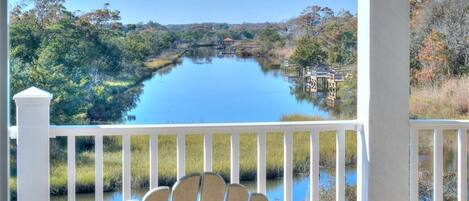 A view from a porch with a wooden chair, overlooking a calm lake with a bridge and dock, surrounded by trees and vegetation.