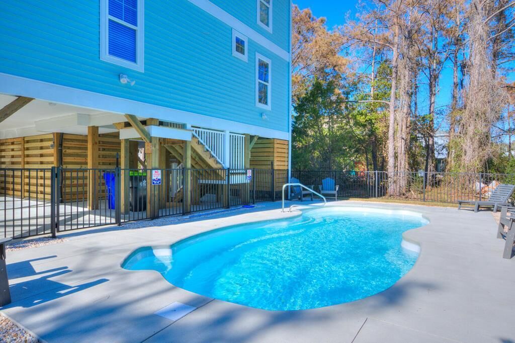 A swimming pool with a wooden deck, a covered area with stairs, and a blue building in the background.