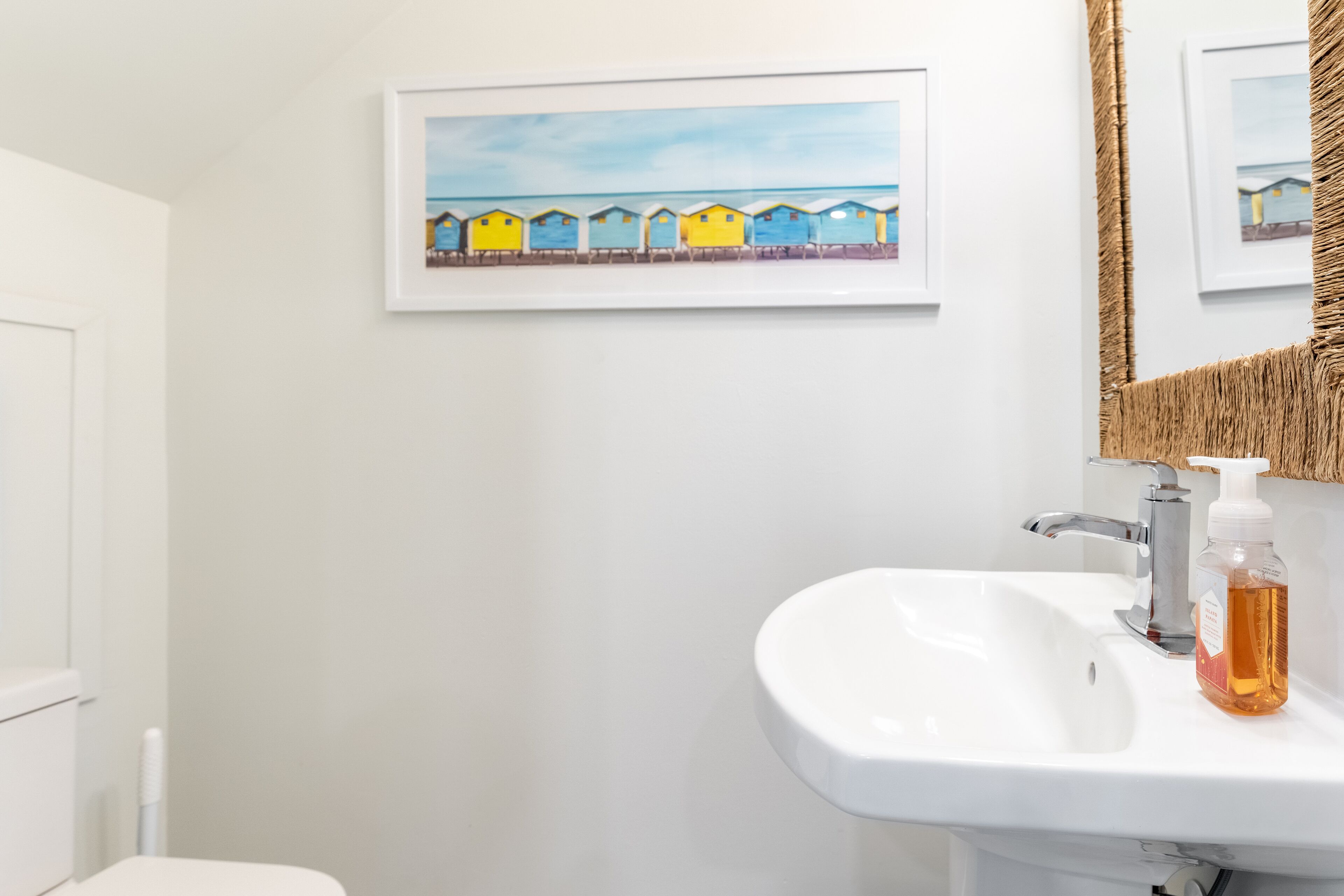 A bathroom with a white sink, a framed painting of colorful beach huts, and a mirror with a woven frame.