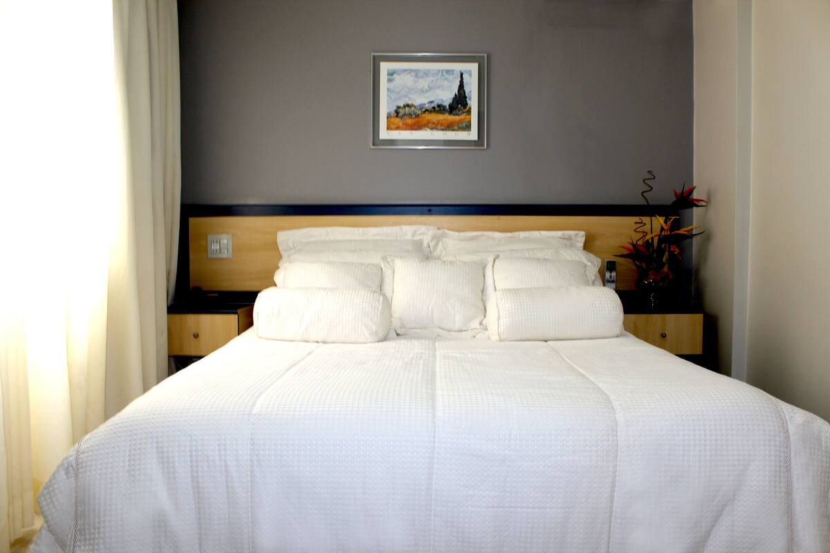 A neatly made bed with a headboard, a framed painting on the wall, and a vase with flowers on the nightstand.