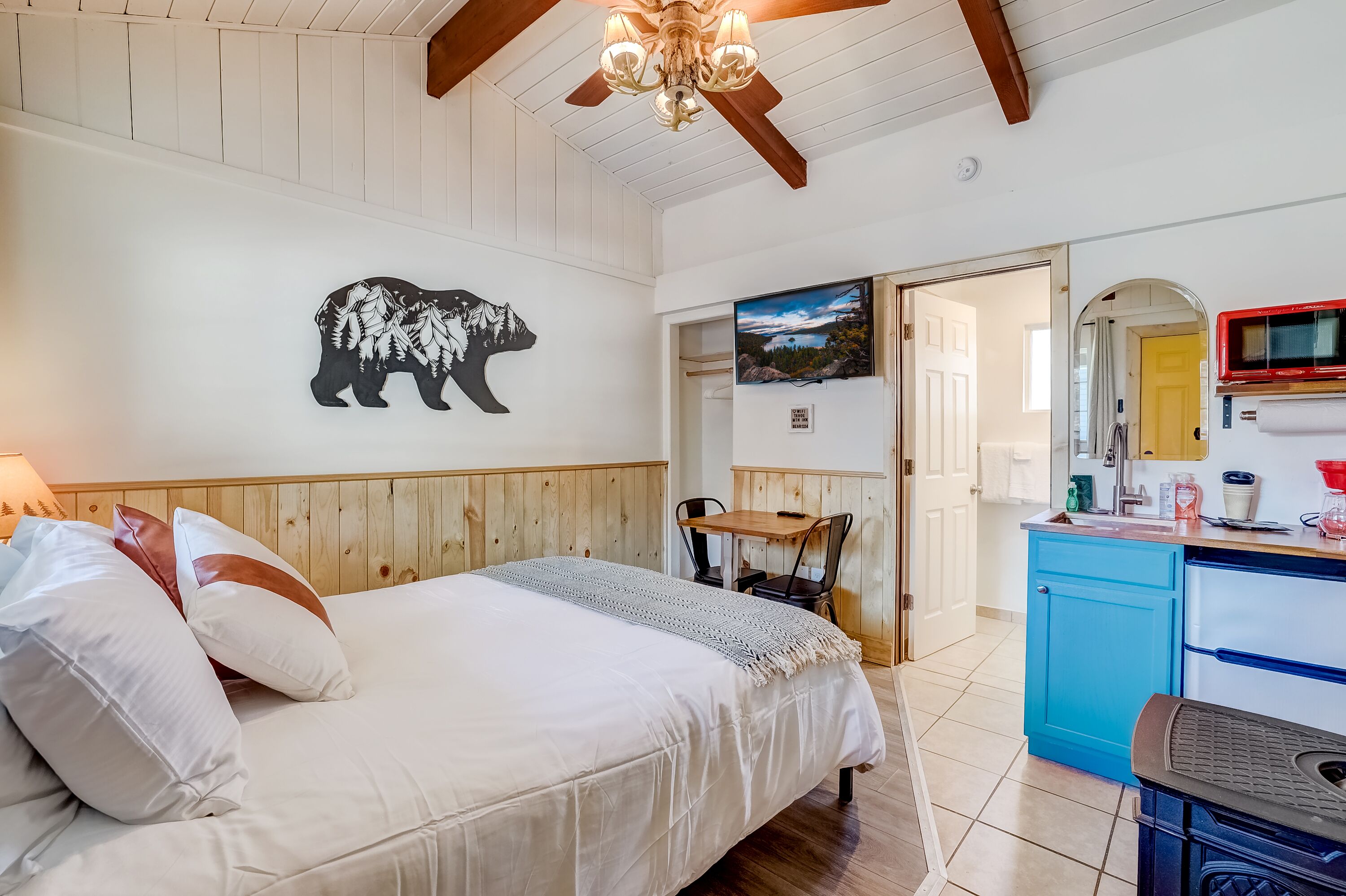 A bedroom with a bed, pillows, a wooden headboard, a wall-mounted bear artwork, a ceiling fan, a kitchen area with a sink, and a microwave.