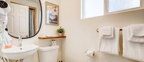 A bathroom with a white sink, toilet, and a large mirror. There is a window above the toilet, a towel rack with towels, and a framed picture on the wall.