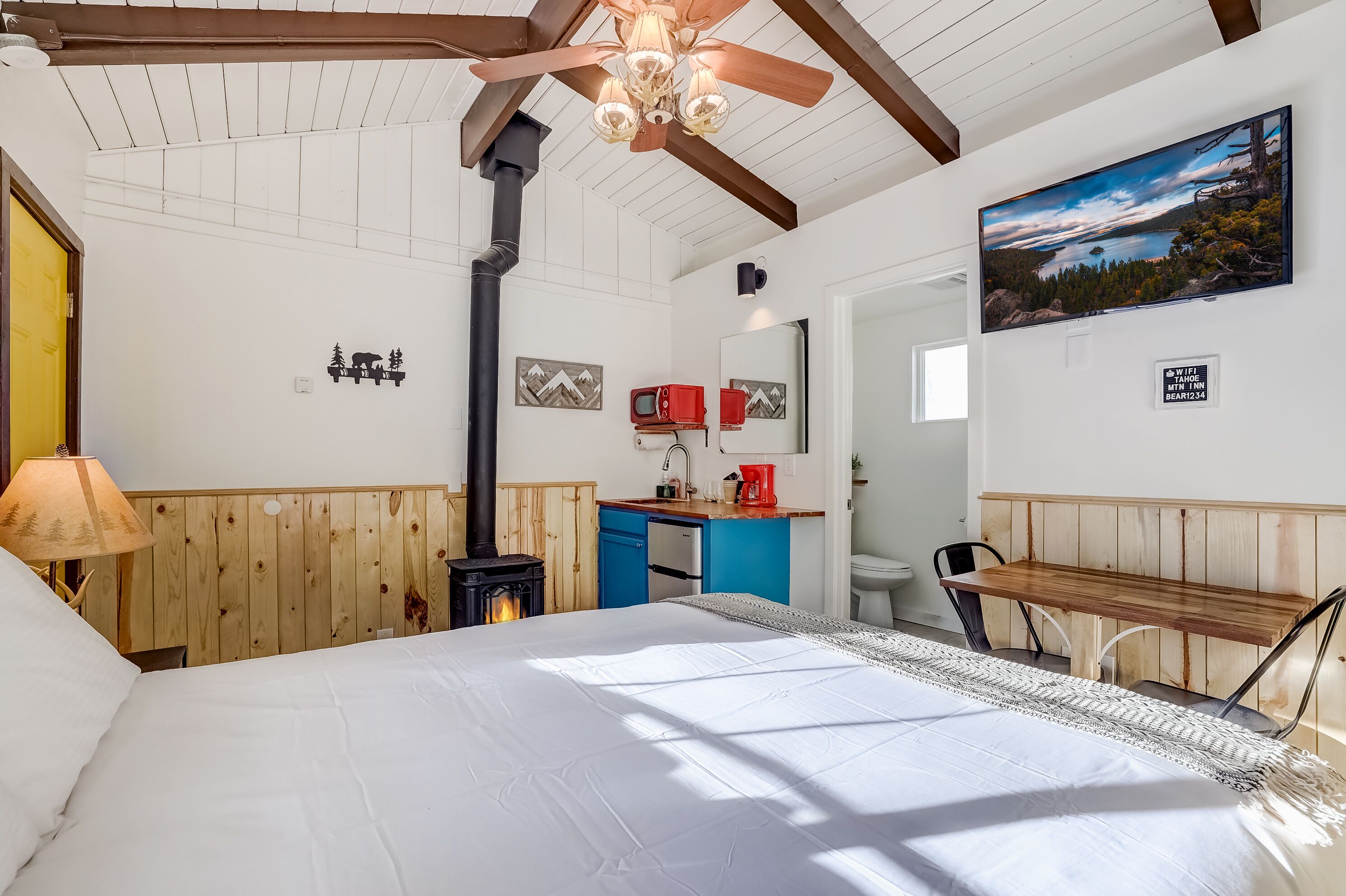 Une chambre avec un lit, un banc en bois, un ventilateur de plafond, un téléviseur fixé au mur et un coin cuisine avec une cuisinière.