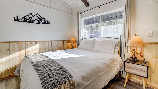 A bedroom with a bed, a nightstand with a lamp, a window with curtains, and a wall-mounted decoration of mountains.