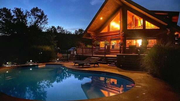 Une cabane en rondins moderne dotée d’une piscine, de chaises longues et d’une terrasse bien aménagée et éclairée.