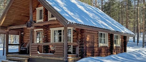 Eine Blockhütte mit einem steilen Satteldach, einer Veranda und verschneiter Landschaft.