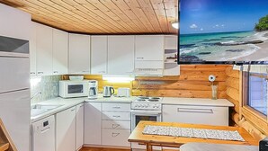 Une cuisine compacte avec des meubles blancs, un four à micro-ondes, une machine à café et une cuisinière. Un téléviseur à écran plat est fixé au mur et diffuse une scène de plage.