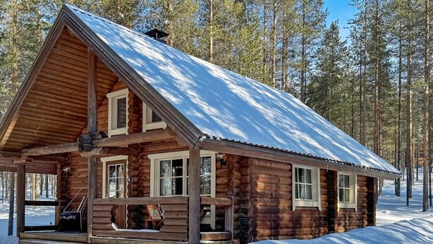 Une cabane en rondins dotée d’un porche couvert et d’un toit enneigé, entourée d’une forêt.