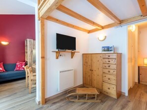 A room with a wooden ceiling, a mounted TV, a wooden cabinet with drawers, a radiator, and a blue sofa with red cushions.