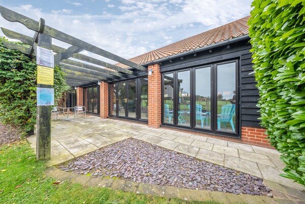 A covered patio area with a stone pathway, a wooden pergola, and a brick building with large windows.