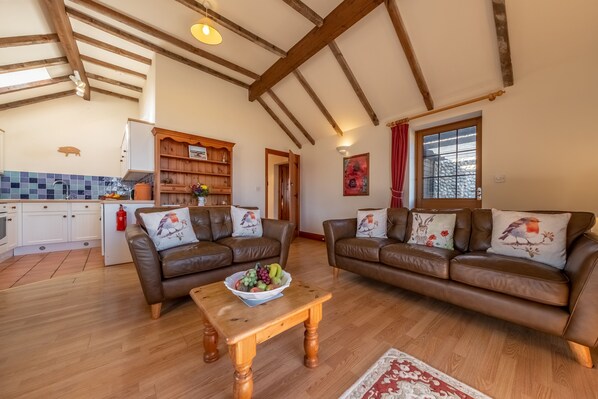 A spacious living area with two brown leather sofas, a wooden coffee table with fruit, a kitchen in the background, and a red curtain on a window.