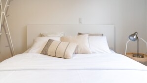 Cama bem arrumada com lençóis brancos e duas almofadas, uma luminária de cabeceira e um interruptor de luz na parede.