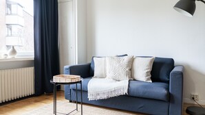 Uma sala de estar moderna com um sofá azul, uma mesinha lateral, um abajur e vista para um edifício.
