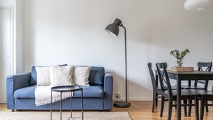 Uma sala de estar com um sofá azul, uma mesa redonda pequena, um abajur e uma mesa de jantar com cadeiras.