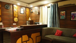 A reception area with a wooden wall, a clock, a desk, a sofa, and a fan. - Hotel J. Palace (New Delhi)