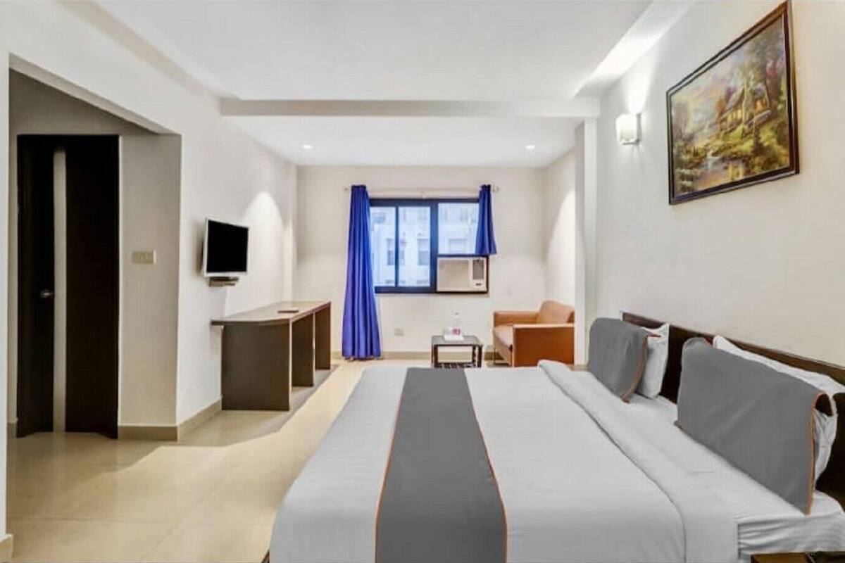 A hotel room with a bed, a TV, a desk, a chair, and a painting on the wall.
