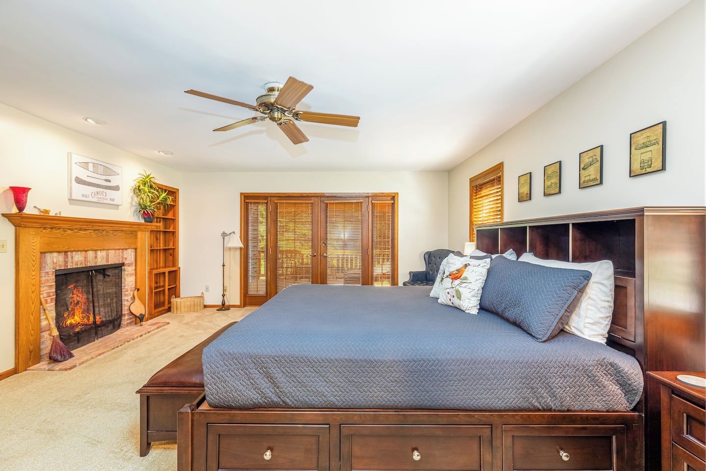 A bedroom with a large bed, a ceiling fan, a fireplace, and framed pictures on the wall.