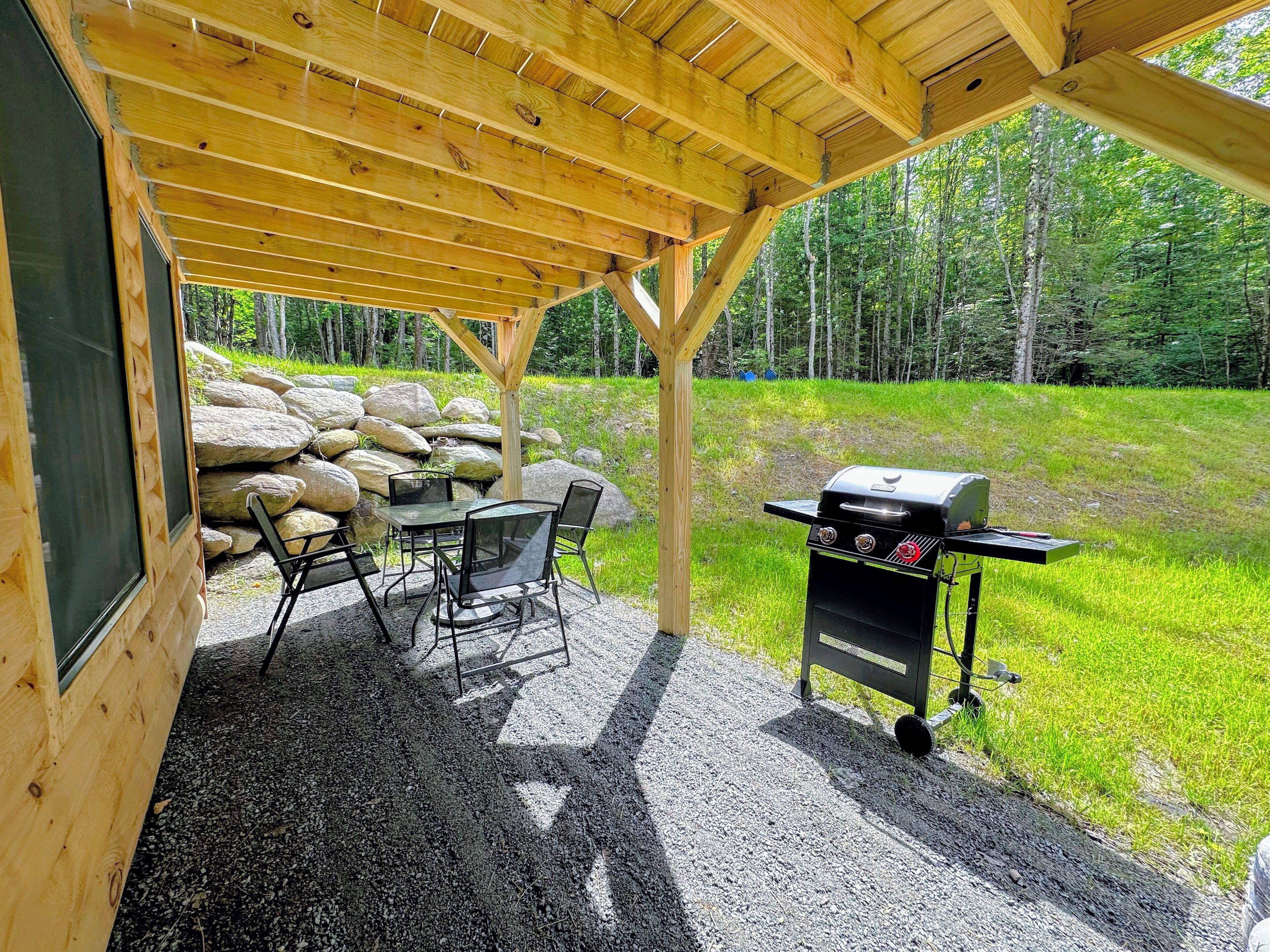 Un espace extérieur abrité, équipé d’une table et de chaises, d’un barbecue et offrant une vue sur une zone boisée.