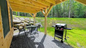 Un espace extérieur abrité, équipé d’une table et de chaises, d’un barbecue et offrant une vue sur une zone boisée.