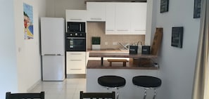 Een moderne keuken met witte kasten, een ingebouwde oven, een magnetron en een koelkast. Er staan twee barkrukken en een bankje bij de toonbank.