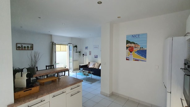Une cuisine moderne avec des meubles blancs, un coin repas avec une table et des chaises, et un salon avec un canapé. Un grand poster est accroché au mur et il y a un réfrigérateur dans la cuisine.