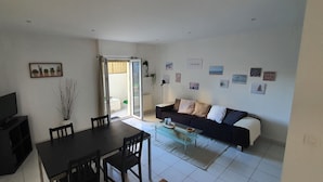 Un salon avec un canapé noir, une table basse en verre, une table à manger avec des chaises, un téléviseur posé sur un meuble et une porte-fenêtre coulissante donnant sur un balcon.