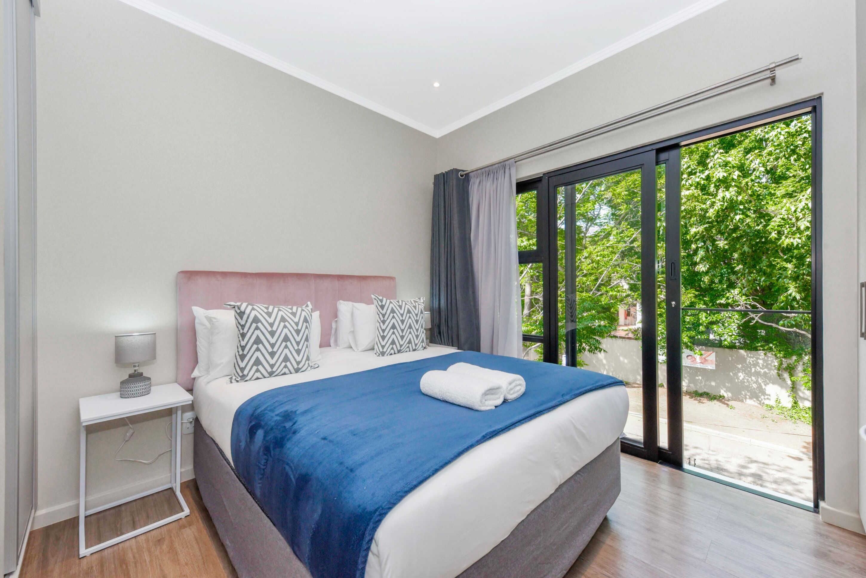 Une chambre moderne avec un grand lit, une table de chevet avec une lampe et une vue sur l’extérieur, accessible par une porte-fenêtre coulissante.