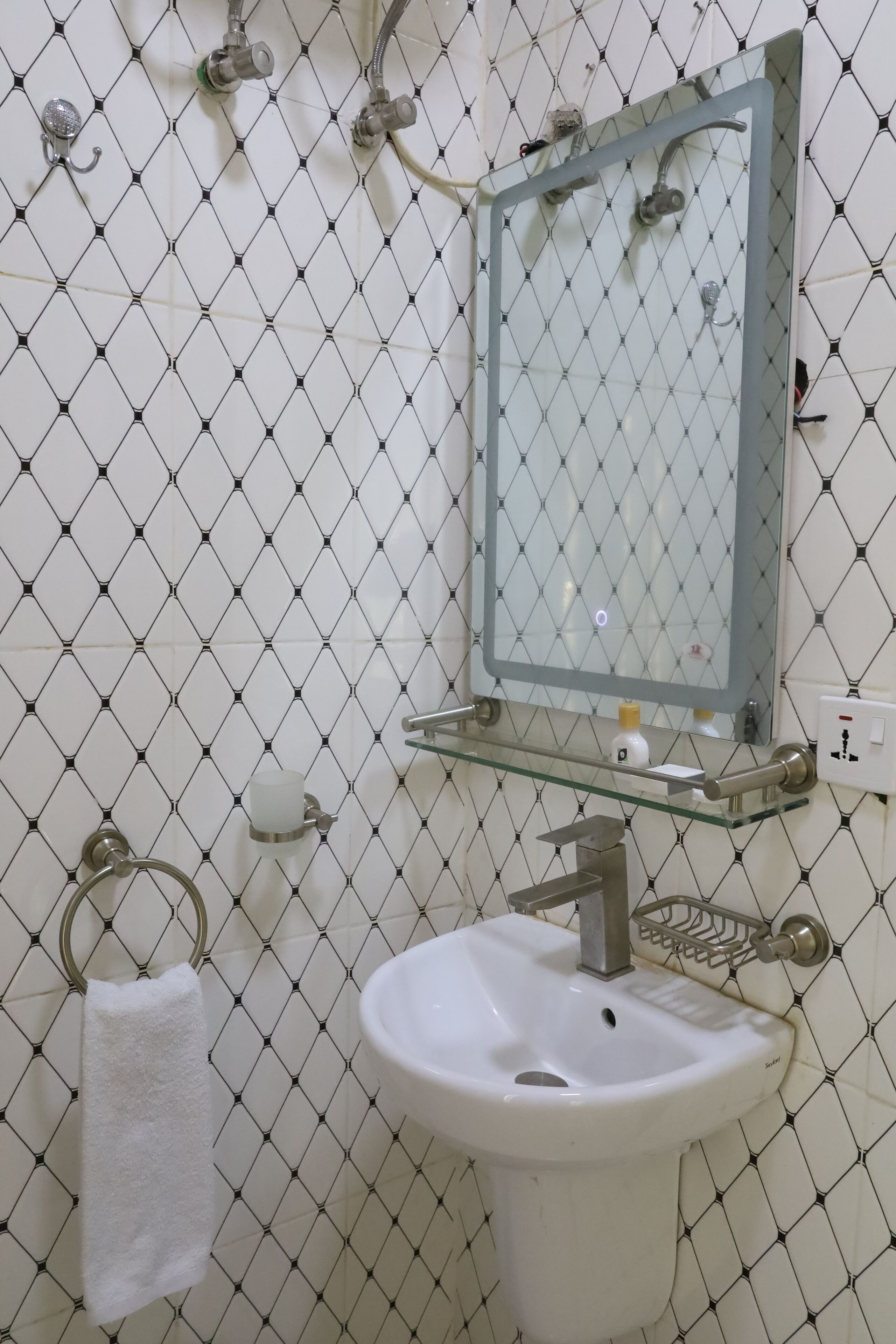 Un baño con pared de azulejos blancos, estante de vidrio sobre un lavamanos blanco, argolla para toallas y un espejo.