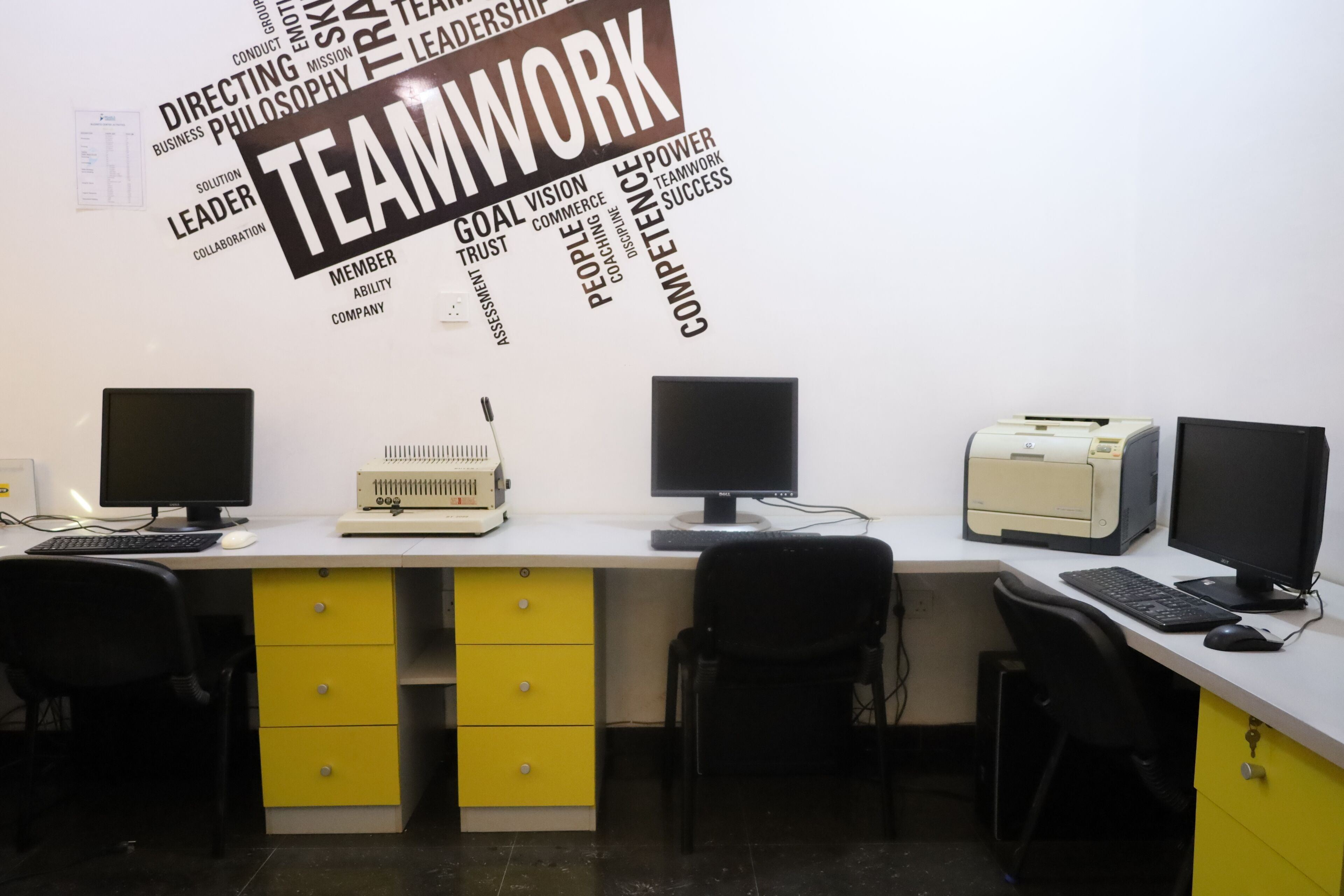 A workspace with two desks, each with a computer monitor, keyboard, and printer. There are yellow drawers under the desks and black office chairs.