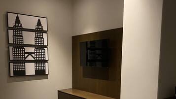 Moderni hotellihuone, jossa on suuri sänky, puinen yöpöytä, seinälle asennettu televisio ja kaupunkimaisemaa esittävä taideteos.