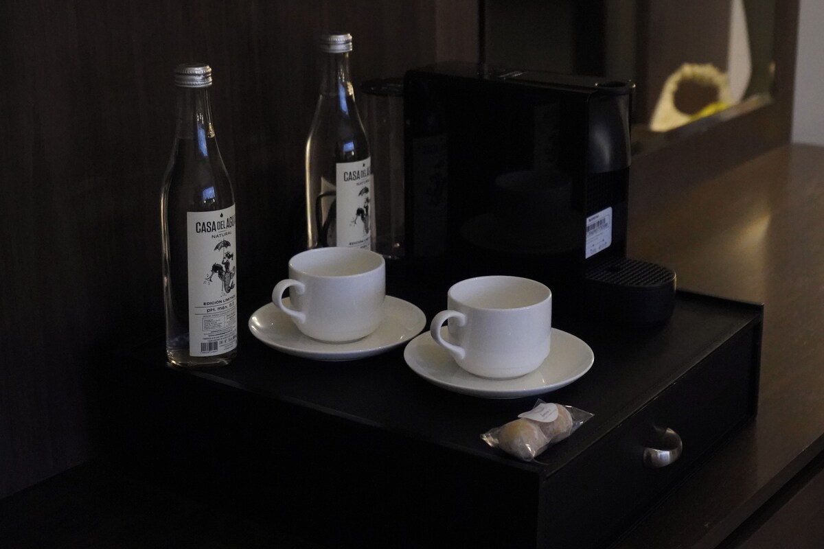 A hotel room's minibar with two white cups, two bottles of water, a coffee maker, and a small packet of snacks.