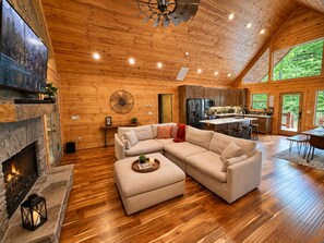 A spacious living room with a stone fireplace, a large sectional sofa, and a dining area with a kitchen in the background. - Hidden Creek Cabin Sleeps 8 • Hot Tub • Game Room • 8 Min to Downtown (Blue Ridge)