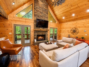 A cozy cabin interior with a stone fireplace, wooden walls, a large sofa, and a television.
