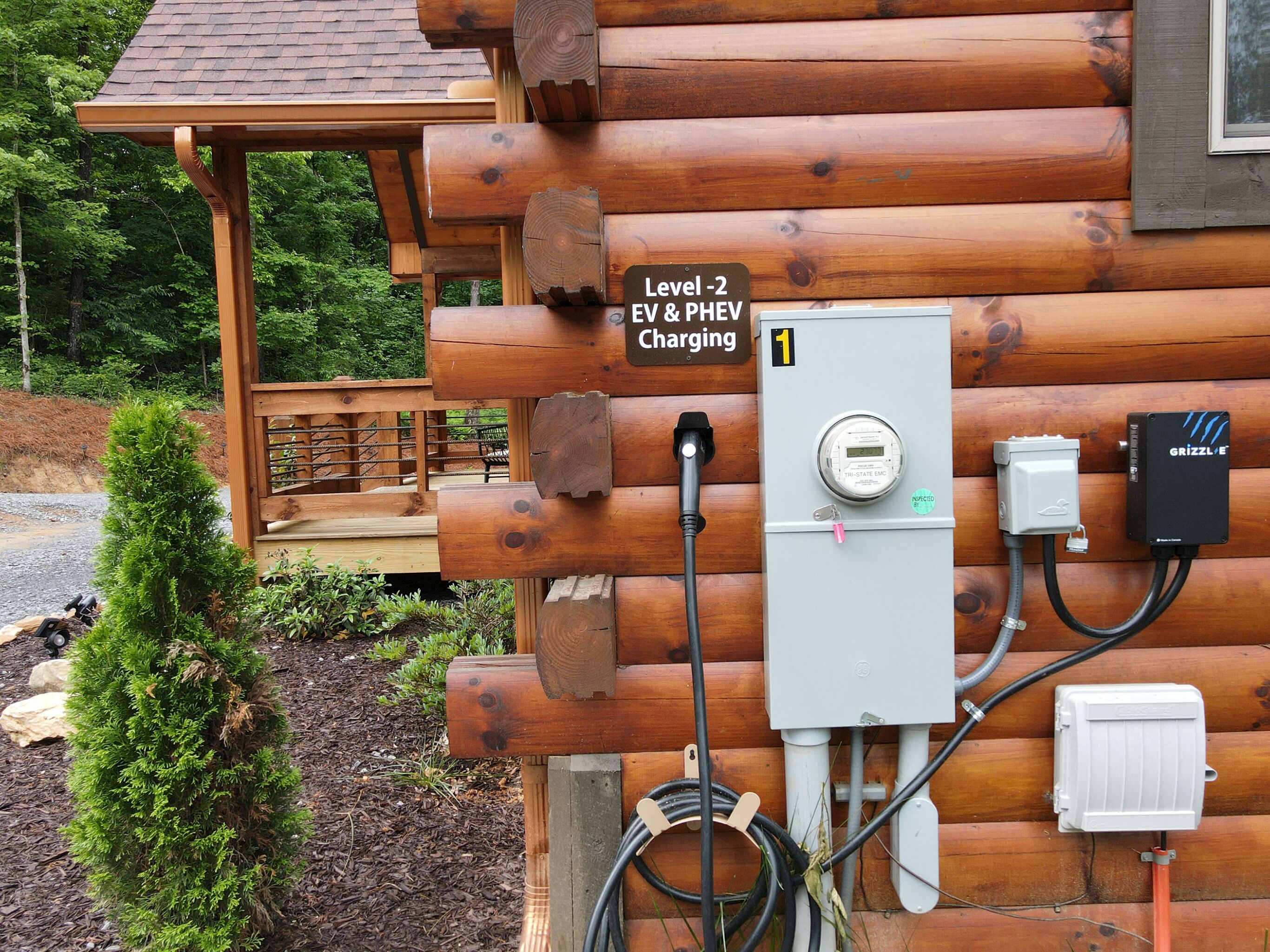 A wooden cabin with a Level-2 EV & PHEV charging station, a small deck, and a surrounding forest.